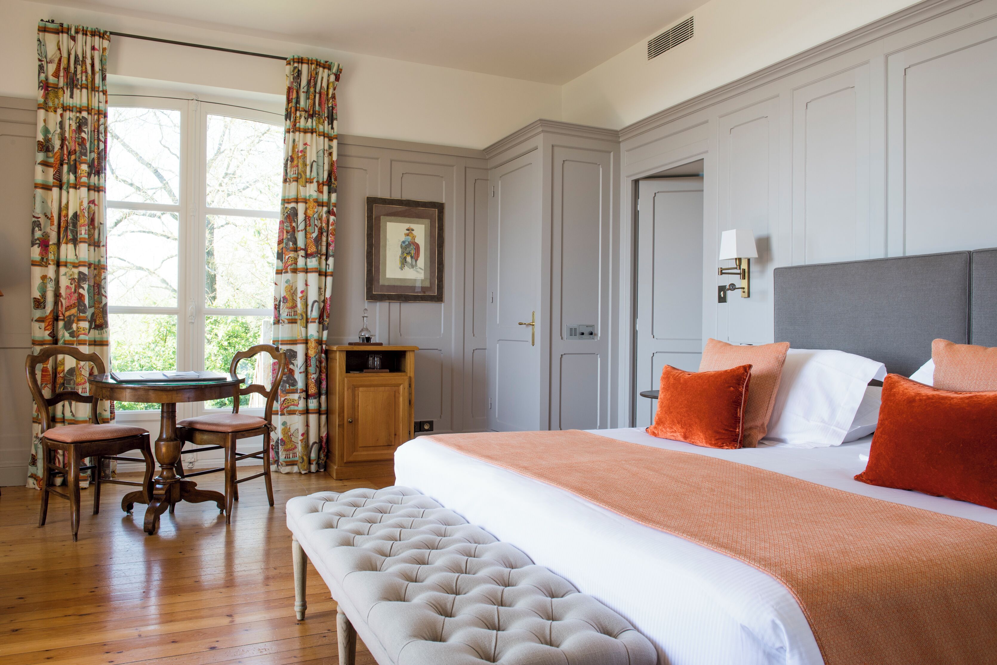 A hotel room with a large bed, a bench, a desk with a chair, and a window with curtains.
