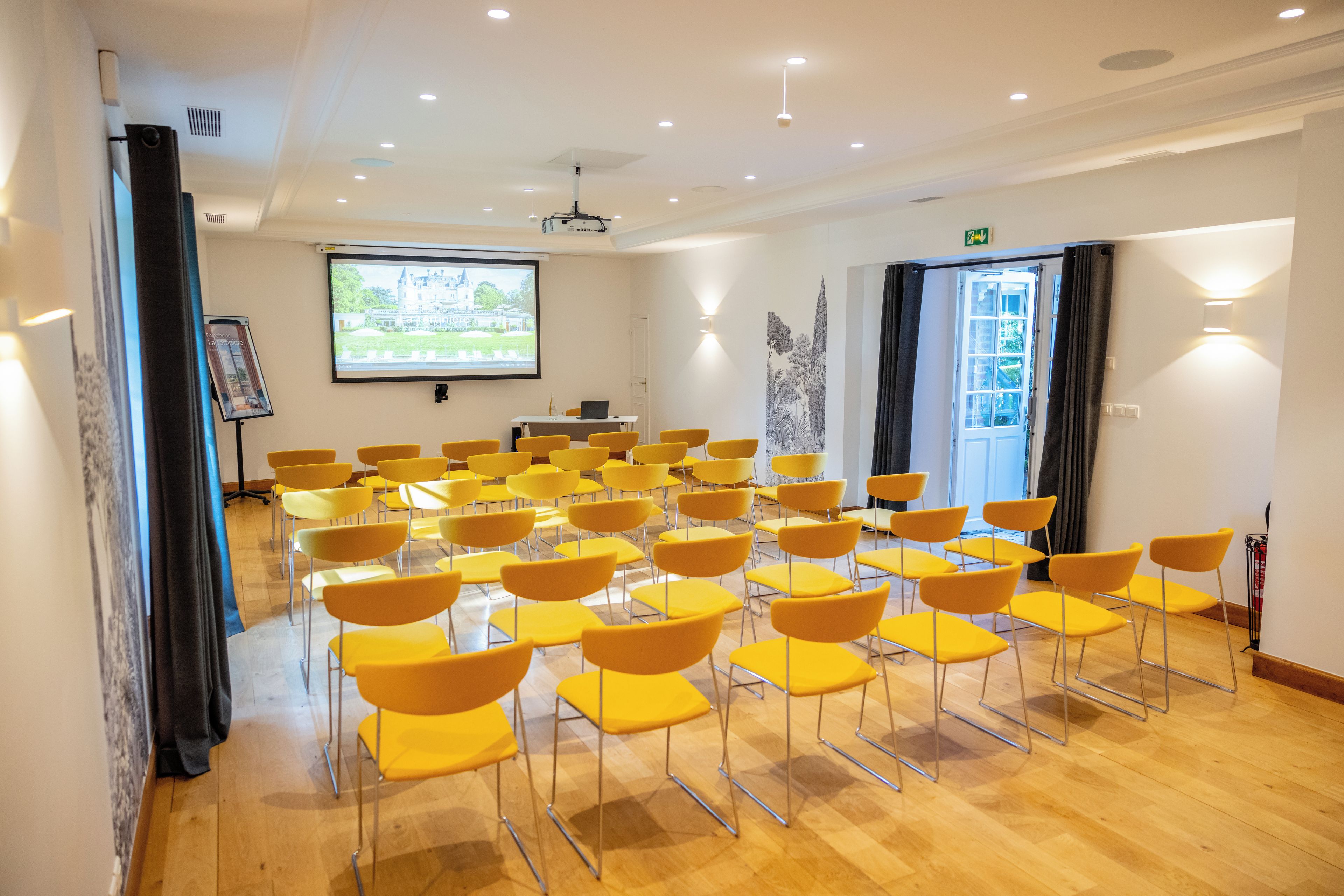 A conference room with rows of yellow chairs, a projection screen, and a door with curtains.