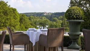 Une table à manger, agrémentée de chaises et d’un vase rempli de verdure, offre une vue sur un paysage luxuriant, avec un bâtiment visible au loin.