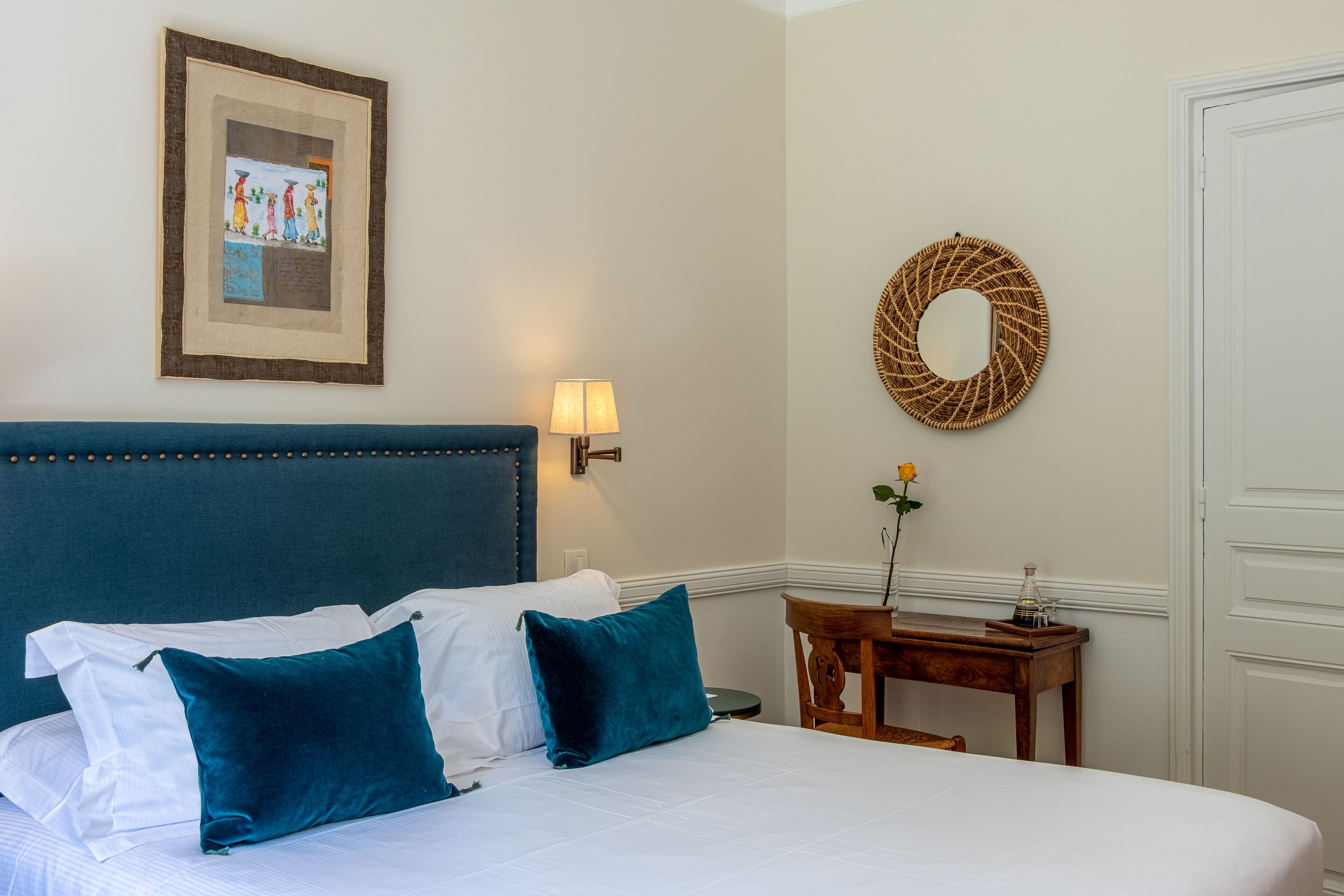 A neatly made bed with two pillows, a wall-mounted picture, a lamp, a mirror, and a small table with a vase.