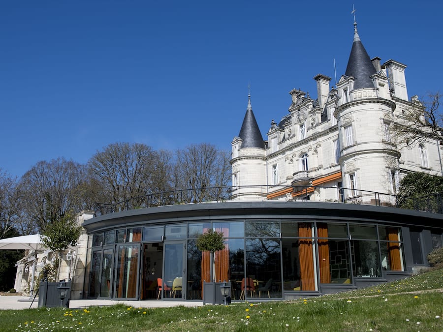 Un edificio imponente con una extensión moderna de vidrio, que presenta un comedor con sillas y mesas, y un gran árbol en primer plano.