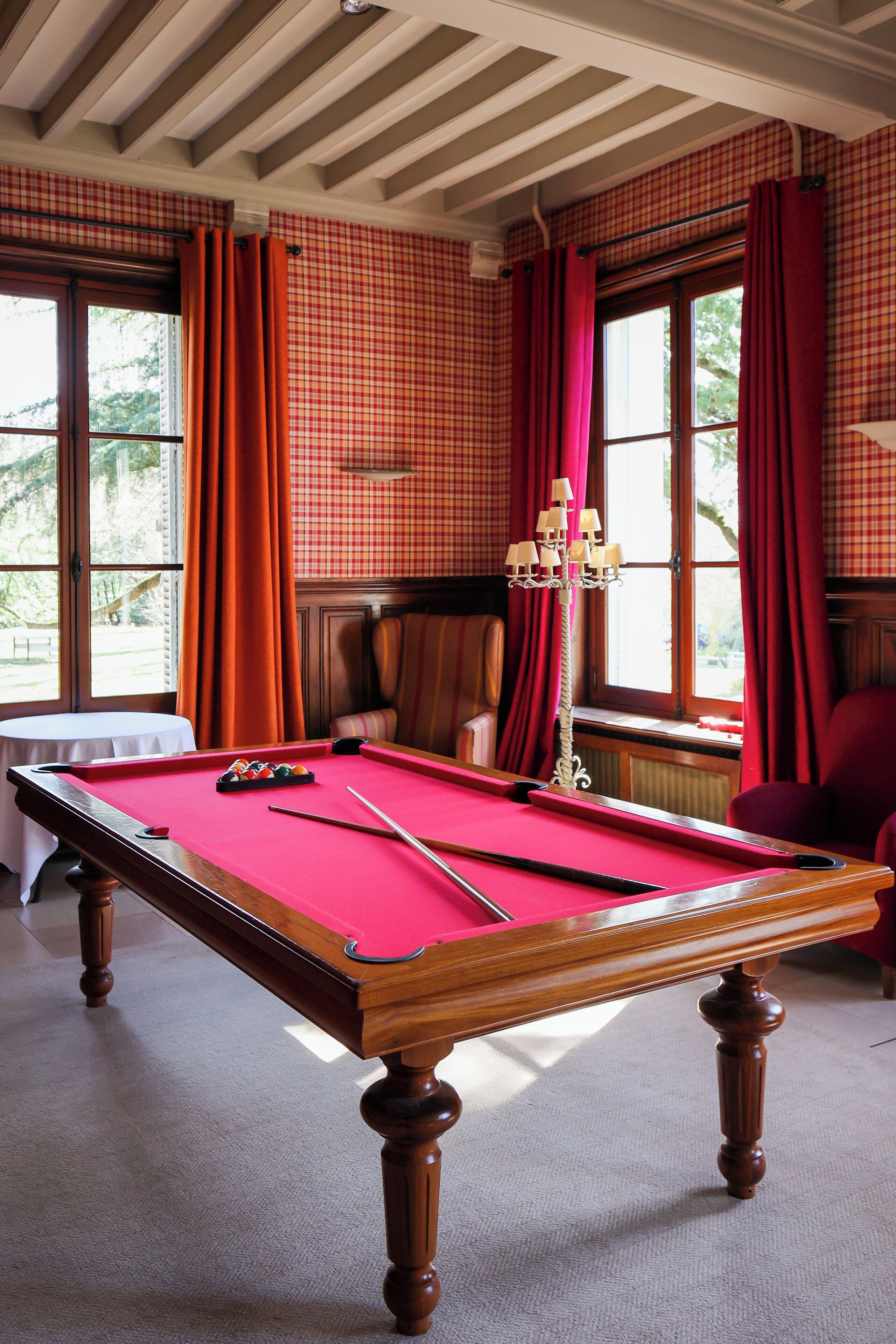 Una mesa de billar en una habitación con papel tapiz a cuadros, techo de madera y ventanas amplias con cortinas rojas.