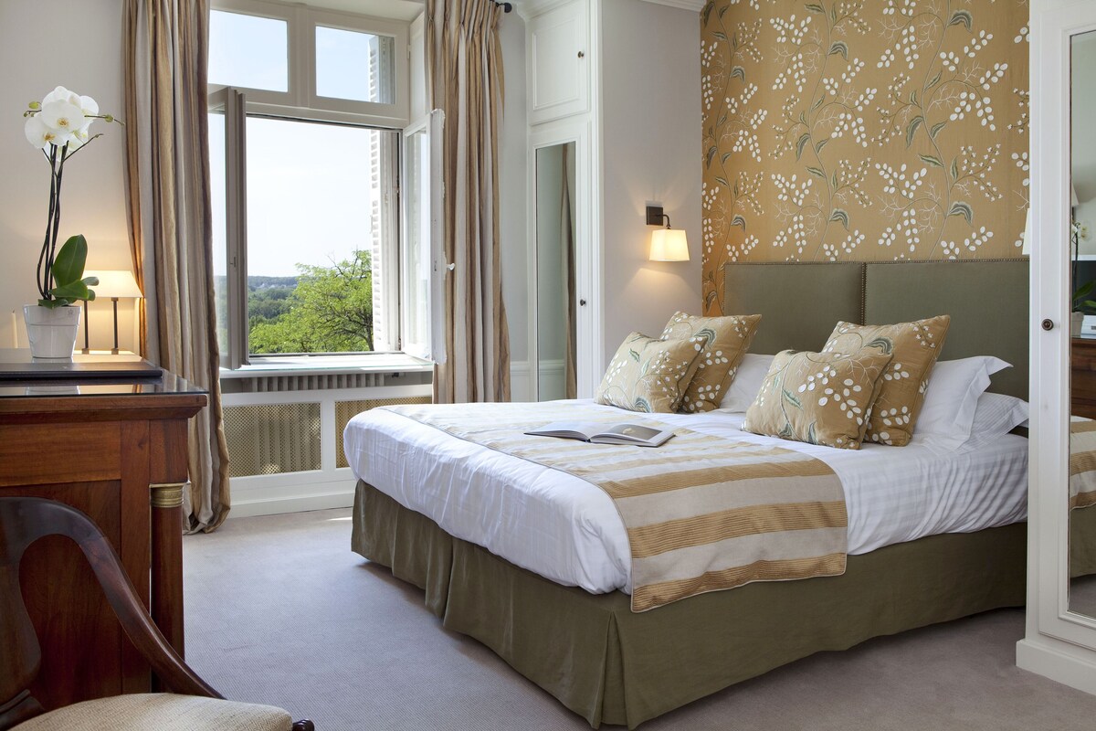 A bedroom with a large bed, a window with curtains, a bedside table with a lamp, and a floral-patterned wall.