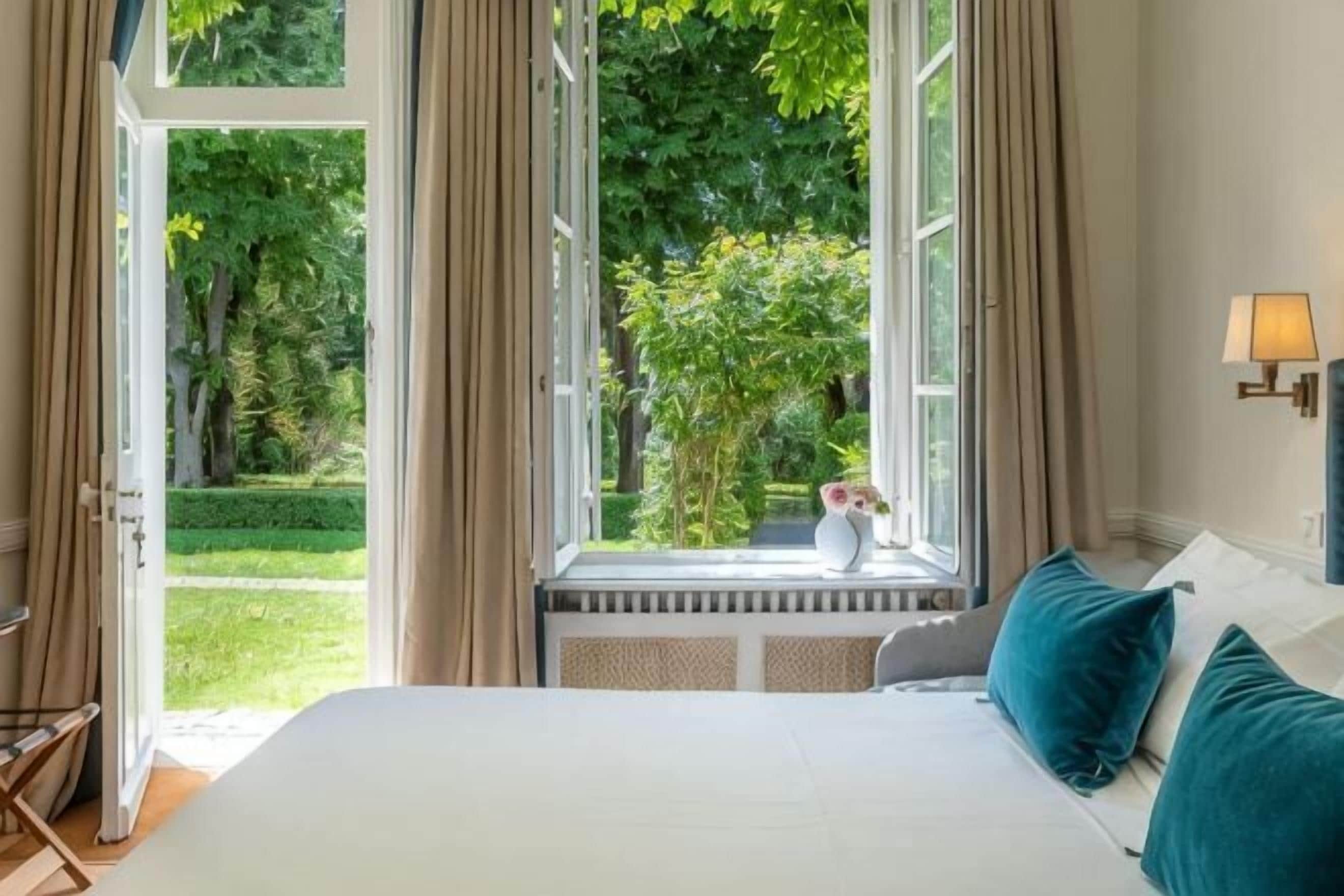 A bedroom with a large bed, a window with curtains, and a view of greenery outside.