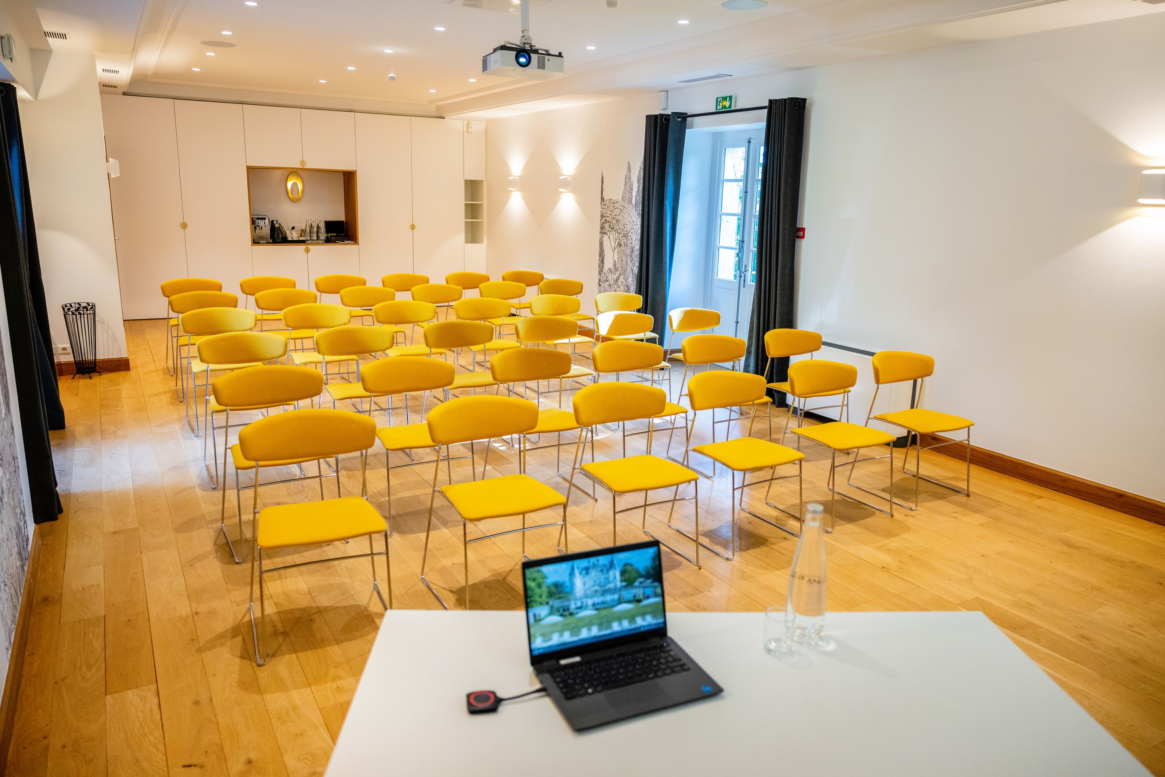 A conference room with rows of yellow chairs, a white table with a laptop, and a projector on the ceiling.