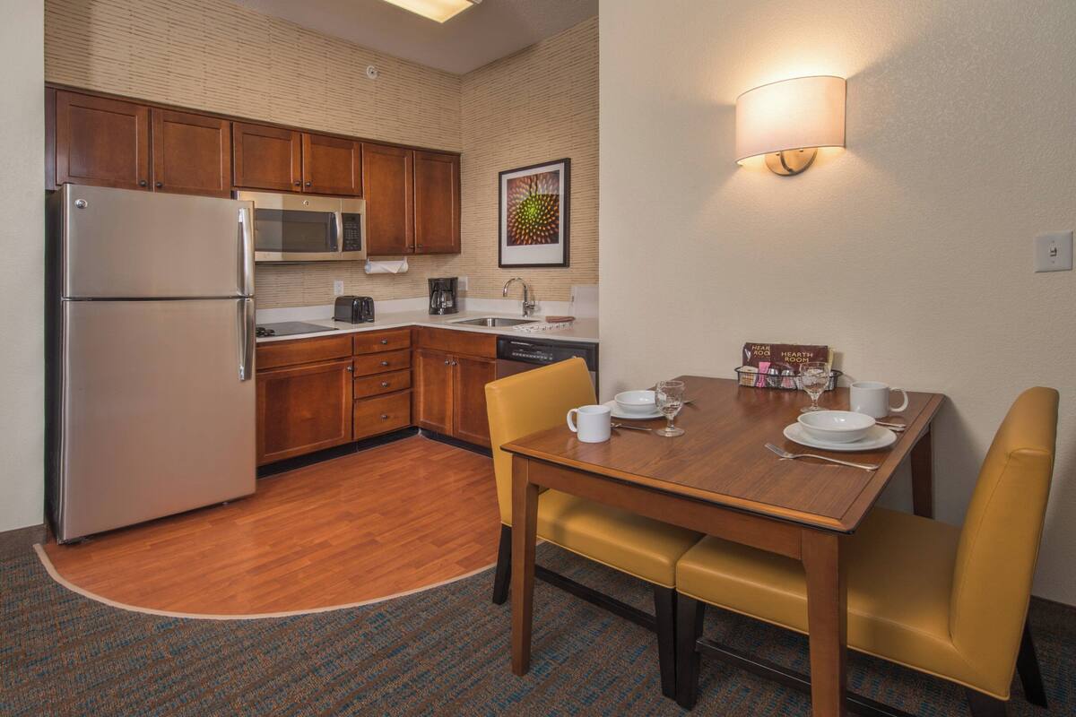 Una cocina compacta con refrigerador, microondas, pileta y muebles de madera. Hay una mesa de comedor con sillas y una lámpara fijada a la pared.