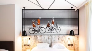 A hotel room with a large bed, two bedside tables, a mounted artwork of people on bicycles, and a wooden accent wall. - ibis Styles Dinan Centre Ville (Dinan)