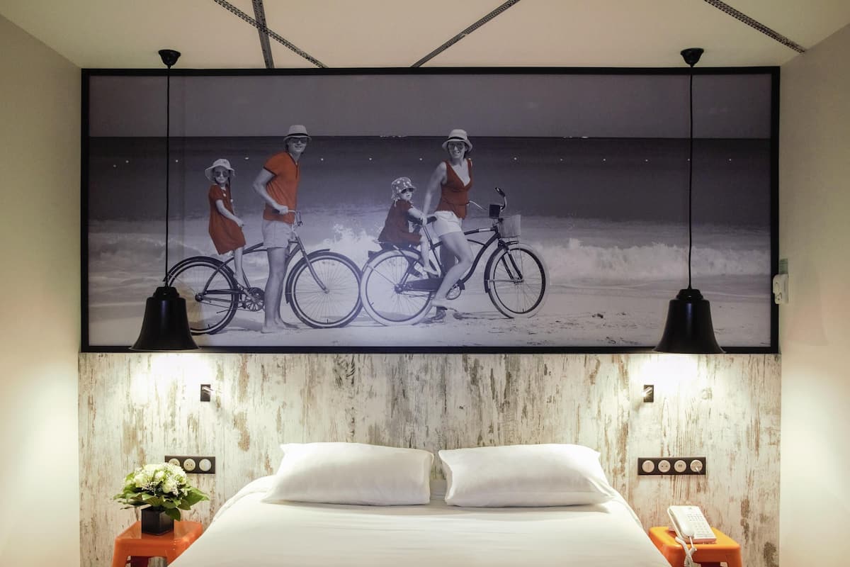 Een slaapkamer met een groot gepresenteerd schilderij van een strandgezicht, een bed met witte lakens, twee nachtkastjes met lampen, en een kleine plant op één nachtkastje.