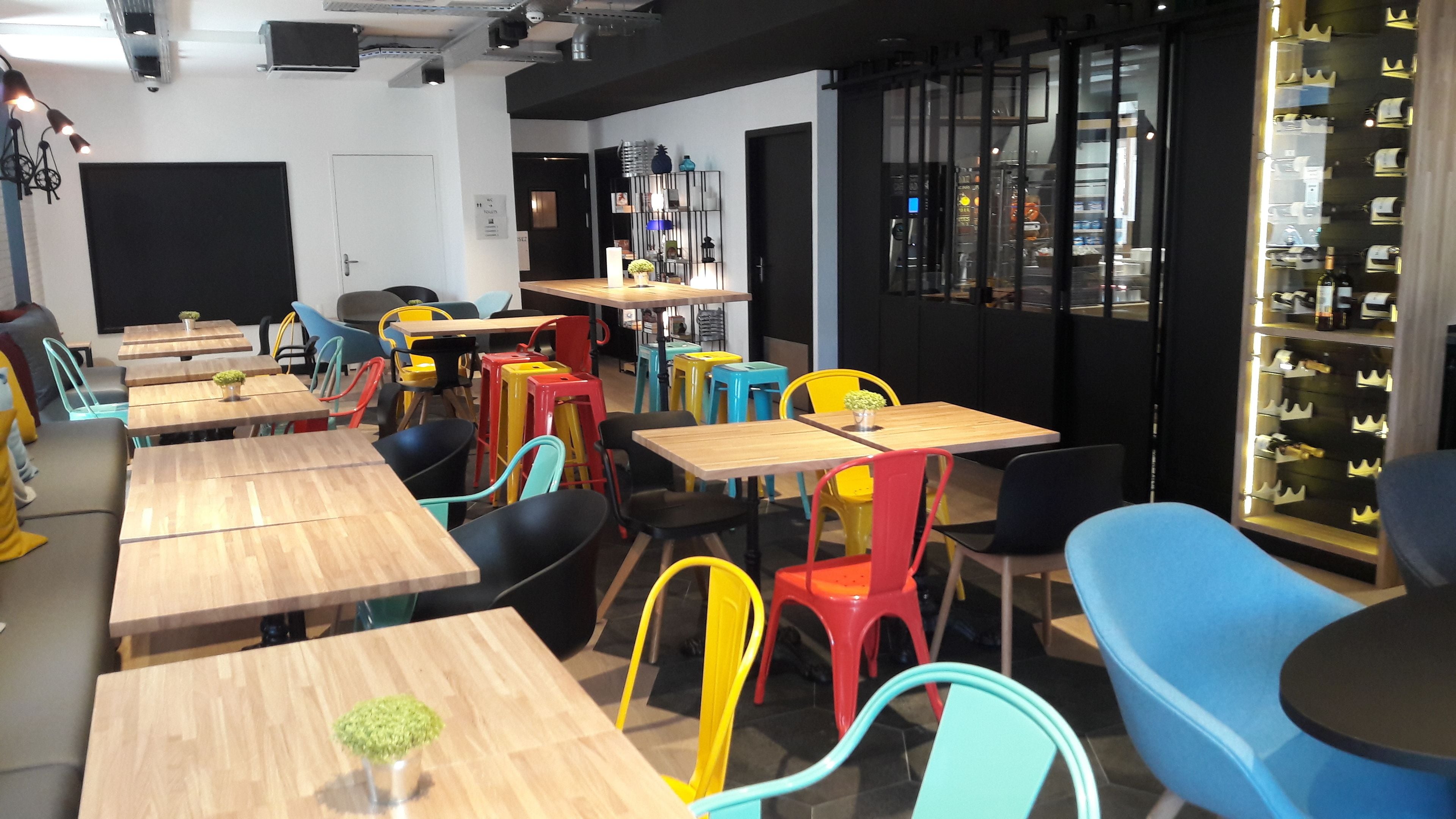 A modern cafe with wooden tables and colorful chairs, a bar area with stools, and a display of bottles.