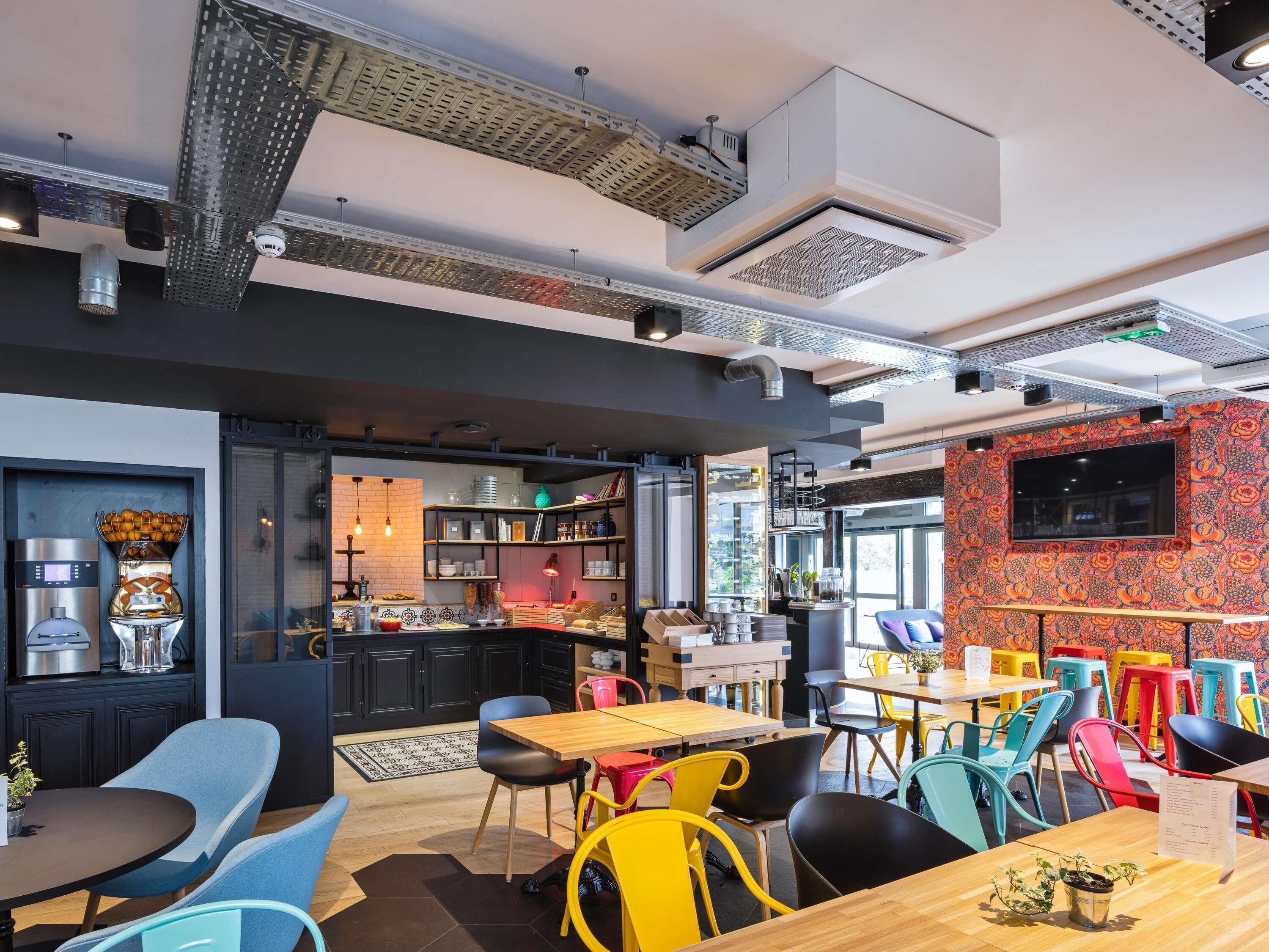 A modern cafe with a kitchen area, colorful chairs, and a large TV screen.