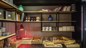 A kitchen area with shelves containing jars, books, and a pineapple. There are also bread baskets and a coffee machine.