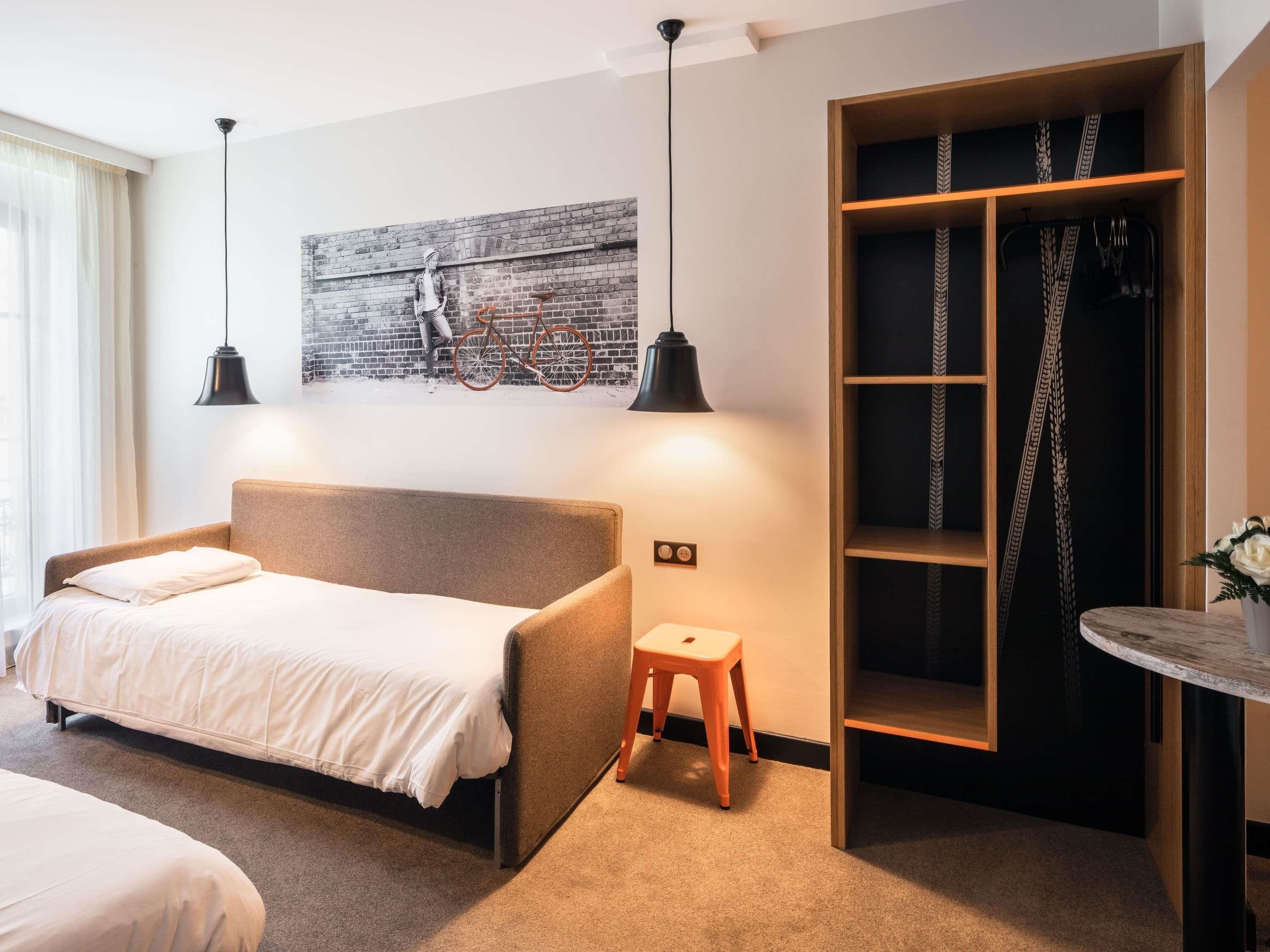 A modern bedroom with a sofa bed, a small stool, a wardrobe with patterned curtains, and a wall-mounted bicycle.