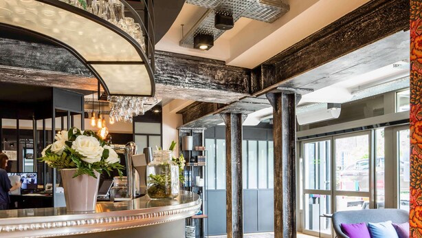 A modern lobby with a curved reception desk, a chandelier, and a seating area with a patterned rug. - ibis Styles Dinan Centre Ville (Dinan)