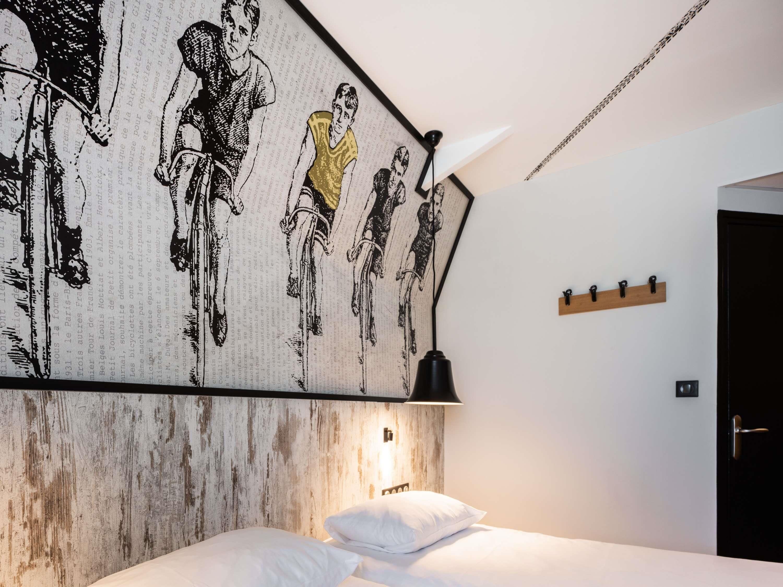 A hotel room with a bed, a wall-mounted lamp, a coat rack, and a framed artwork.