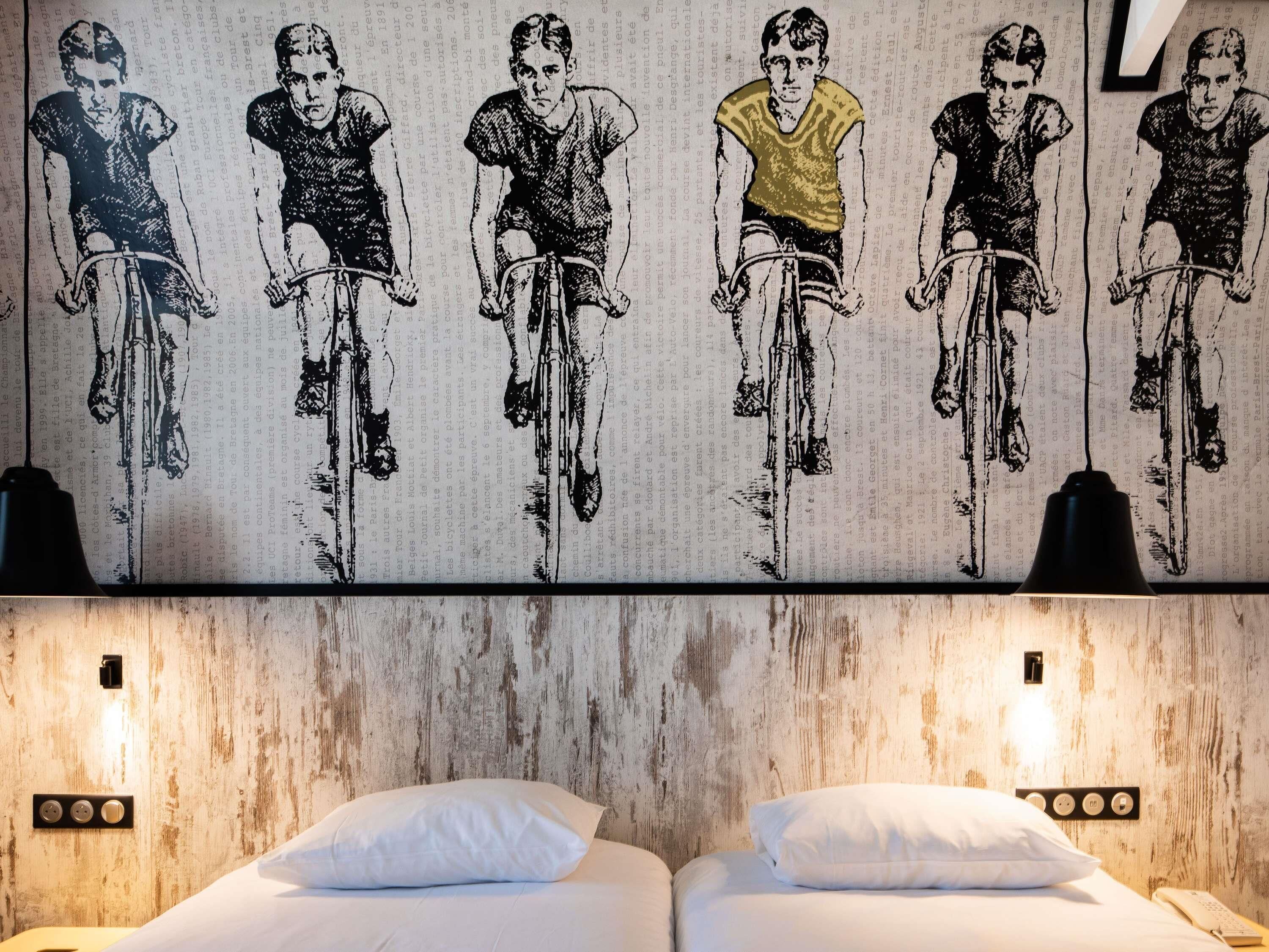 Camera d'albergo con un grande affresco raffigurante ciclisti, due letti con lenzuola bianche e una parete con texture in legno.