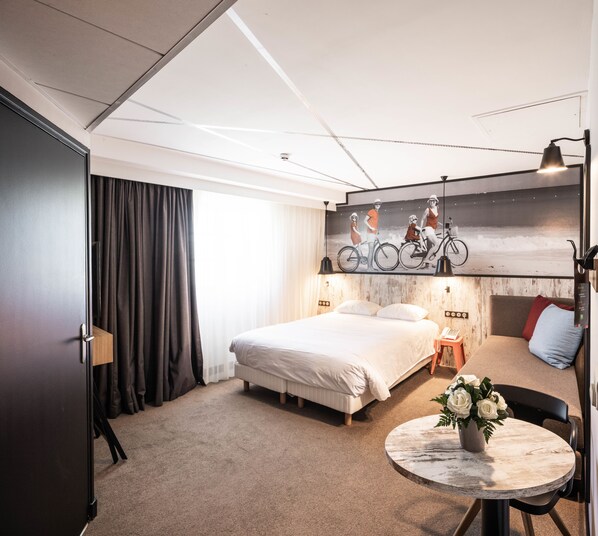 A hotel room with a bed, a small table with a vase of flowers, a desk with a computer, and a wall-mounted artwork of bicycles. - ibis Styles Dinan Centre Ville (Dinan)