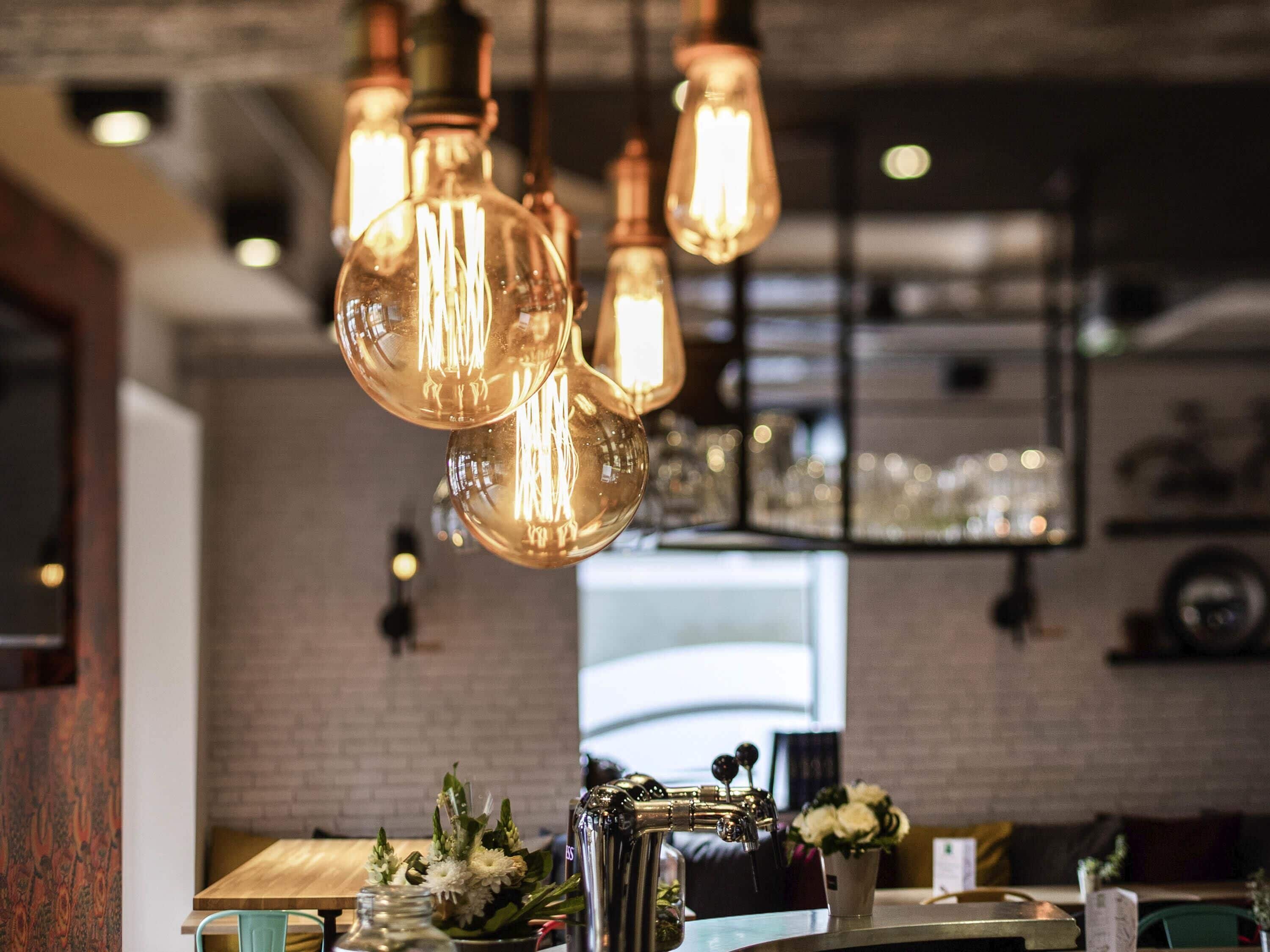 A restaurant interior with hanging Edison bulbs, a brick wall, and a bar area with a tap and flowers.