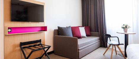 A hotel room with a flat-screen TV mounted on a wooden panel, a sofa with cushions, a small table with a vase, a chair, and a bed.