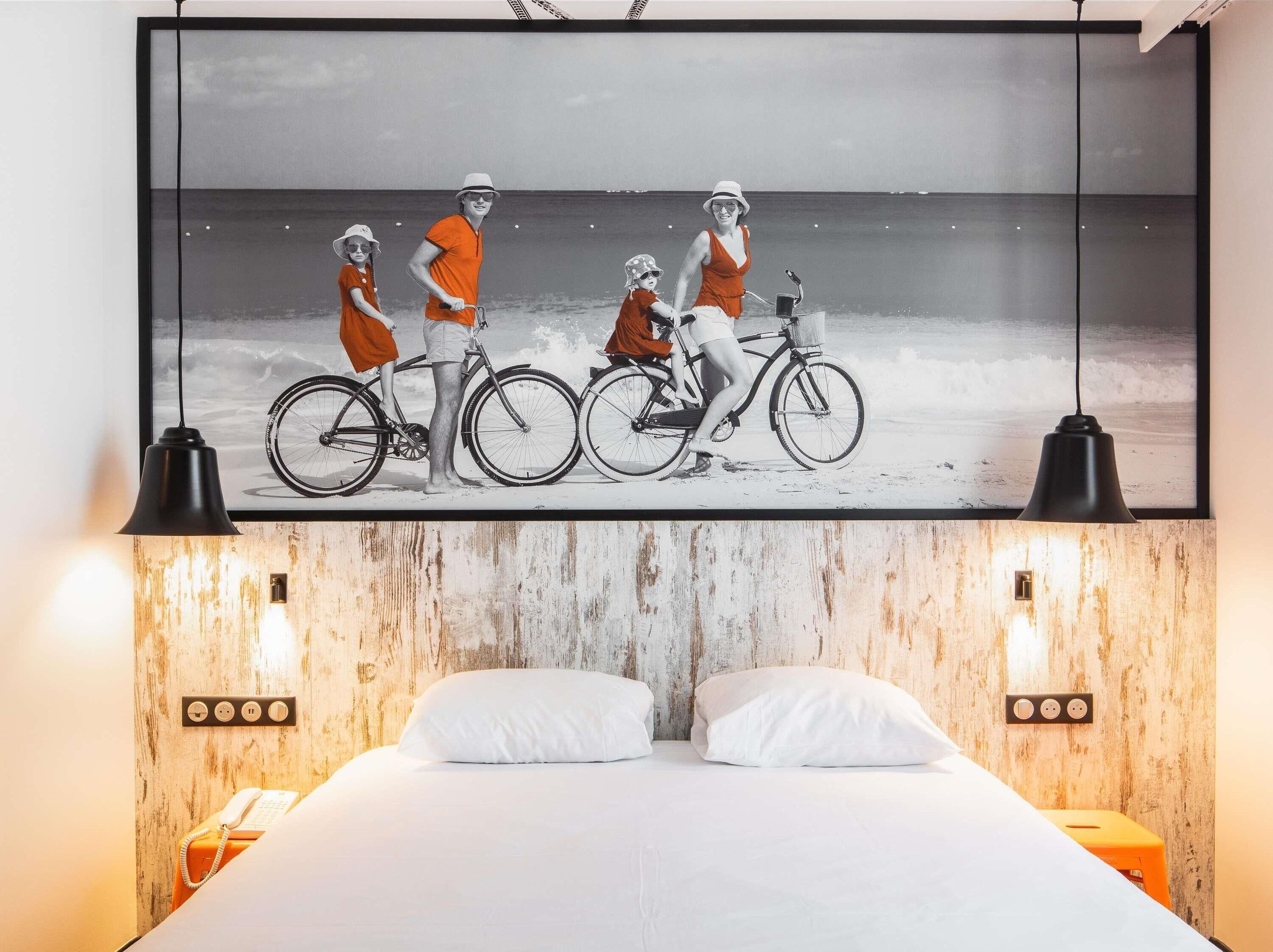 A hotel room with a large framed picture of a beach scene, a bed with white linens, two bedside lamps, and a wooden headboard.