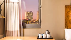 Ett hotellrum med ett skrivbord i trä, en vit stol, en säng, ett fönster med utsikt över staden och en plats för att göra kaffe.