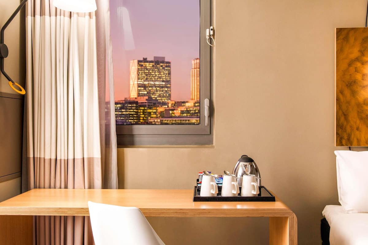 A hotel room with a wooden desk, a white chair, a bed, a window with a cityscape view, and a coffee station.