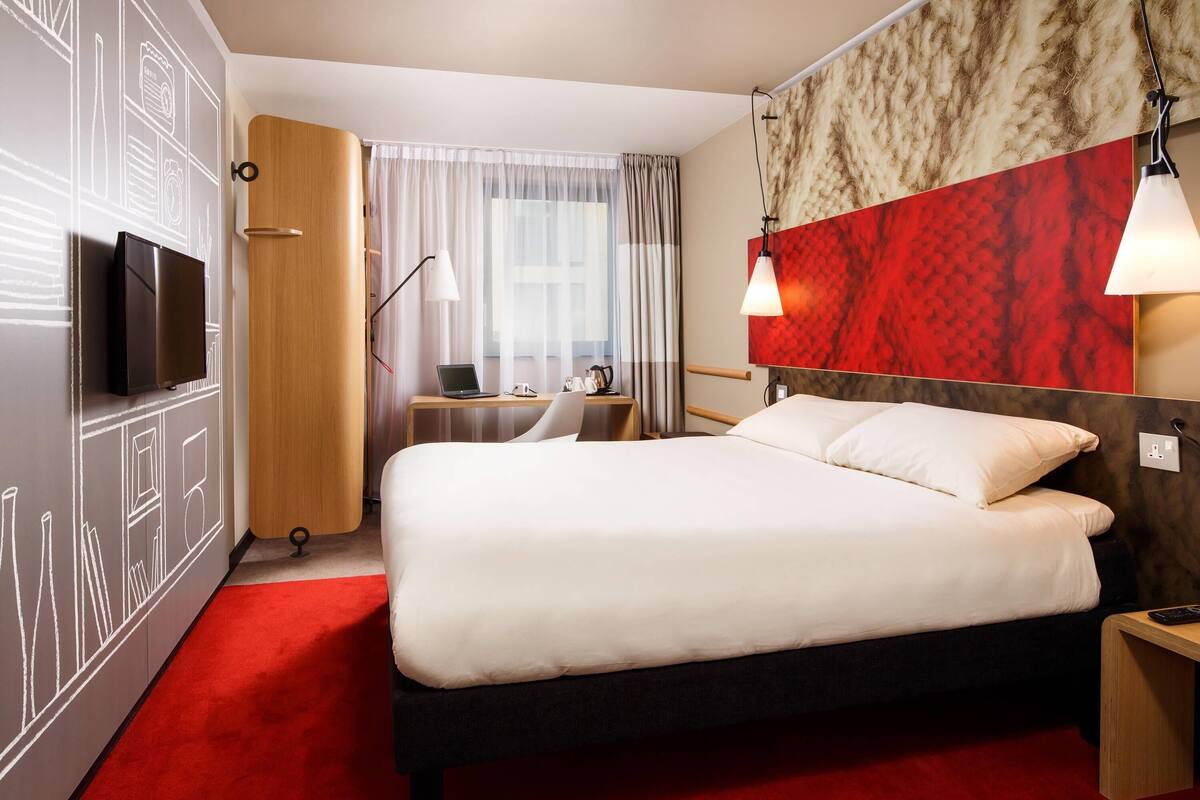 A hotel room with a large bed, a desk with a laptop, a TV, and a red carpet.