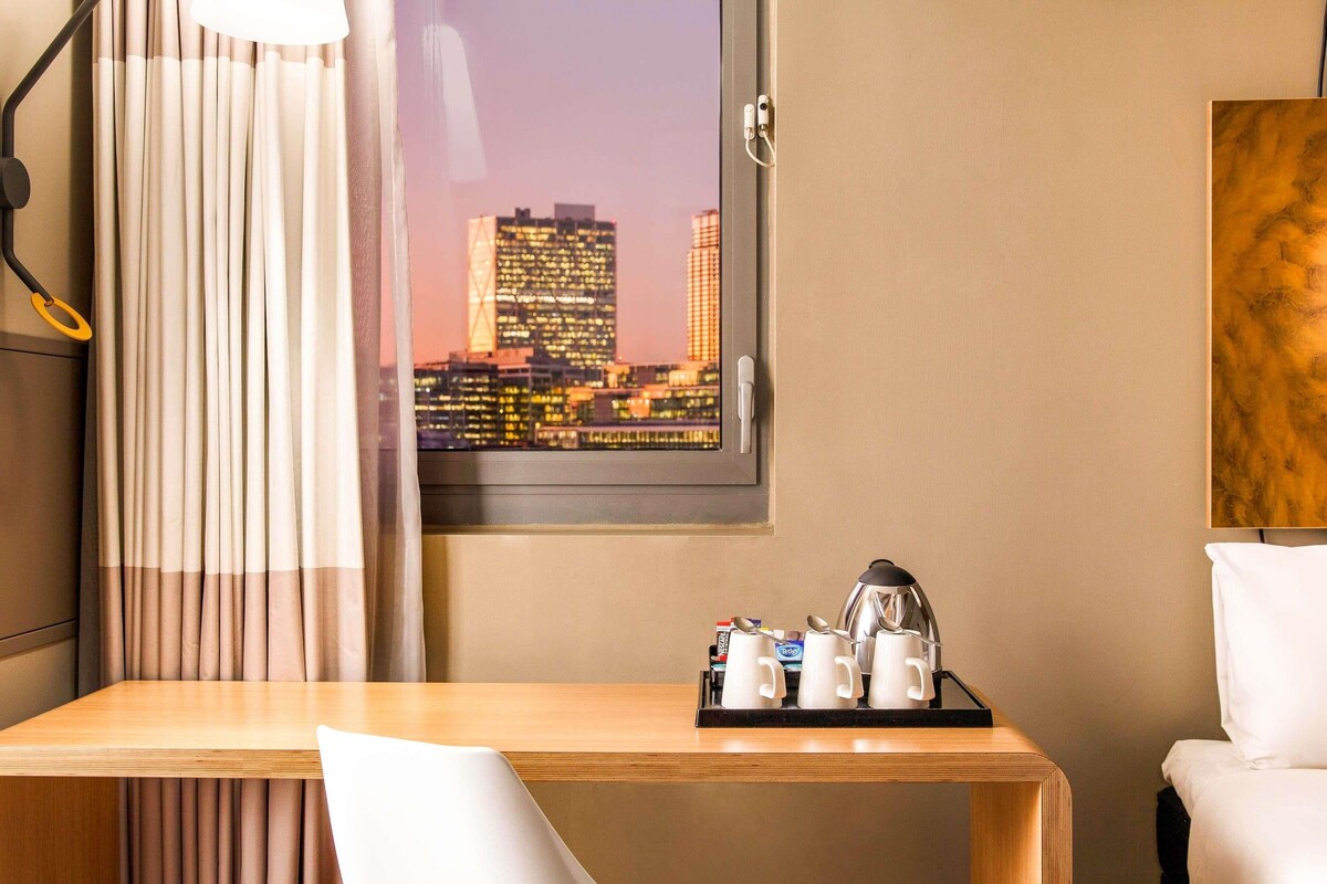 Habitación de hotel con vista a la ciudad, un escritorio de madera, una silla blanca, una cama y una estación de café.