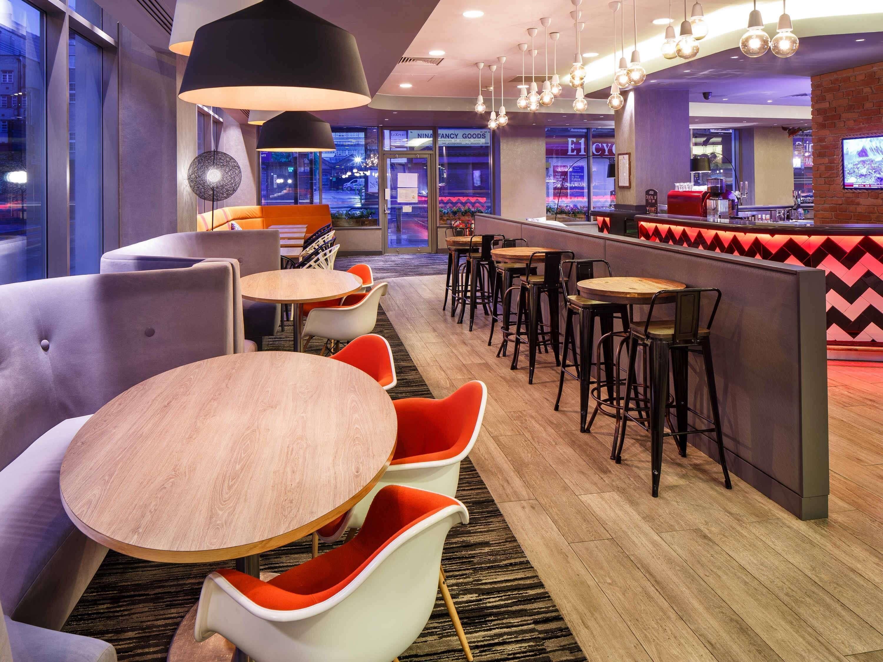Een moderne bar met ronde tafels en barkrukken, een zithoek met gestoffeerde stoelen, en uitzicht op de straat.