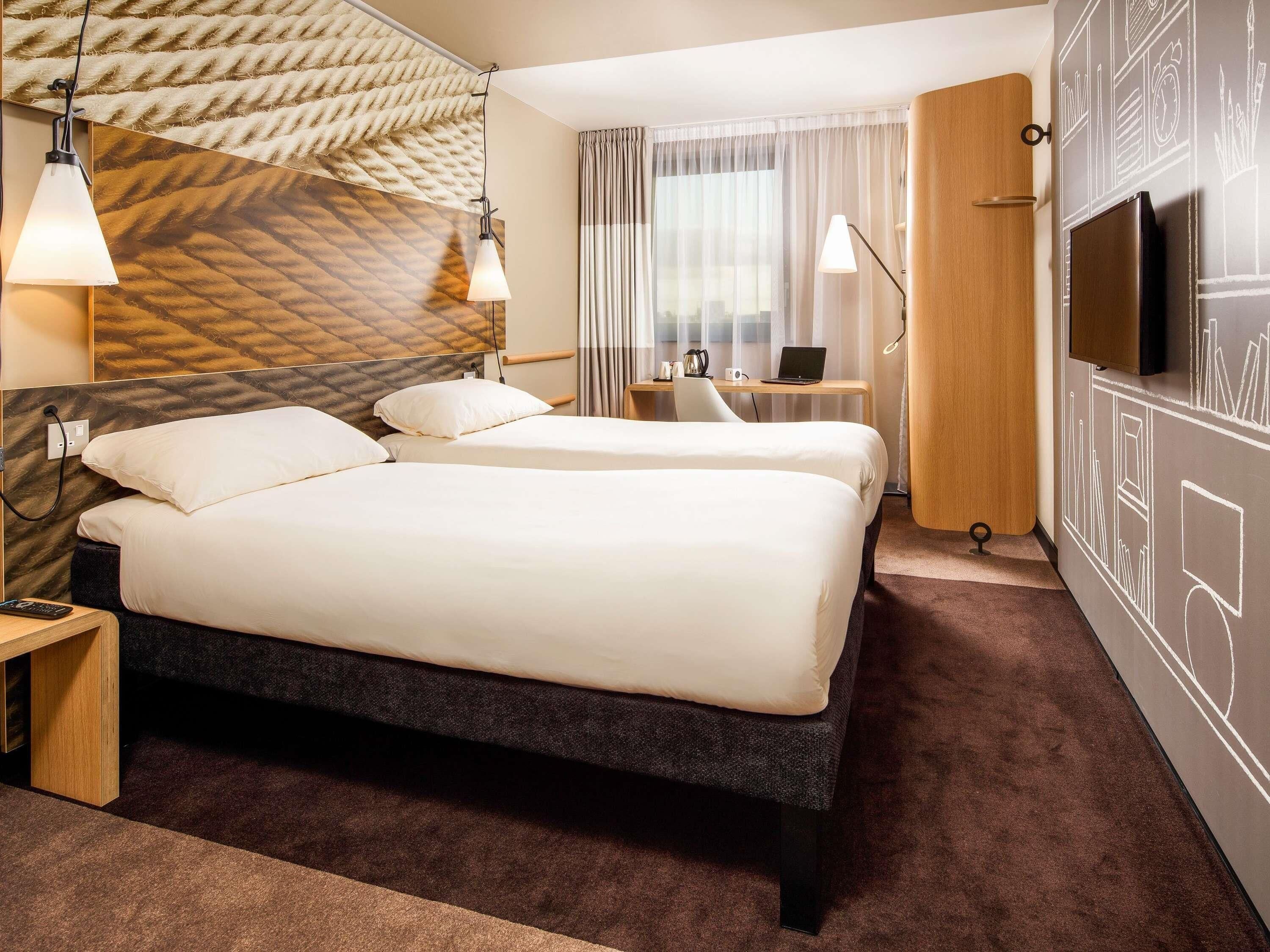 A hotel room with two beds, a desk with a laptop, a TV, and a patterned wall.