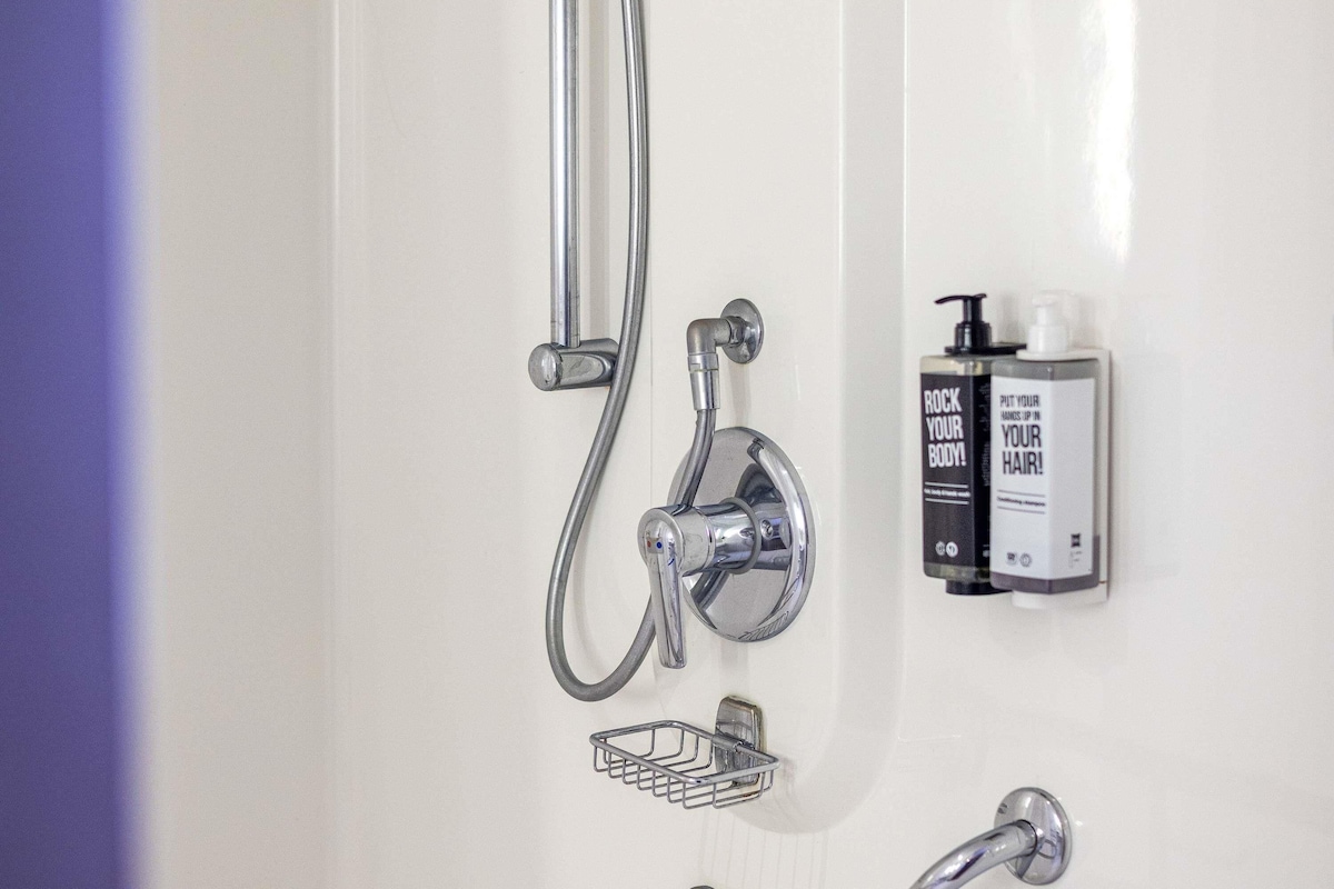 A bathroom with a shower, a soap dispenser, and a small metal basket.