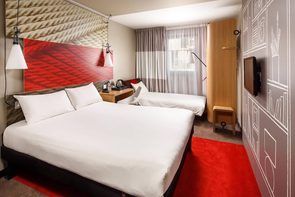 A hotel room with a large bed, a smaller bed, a desk, a chair, a TV, and a red carpet.