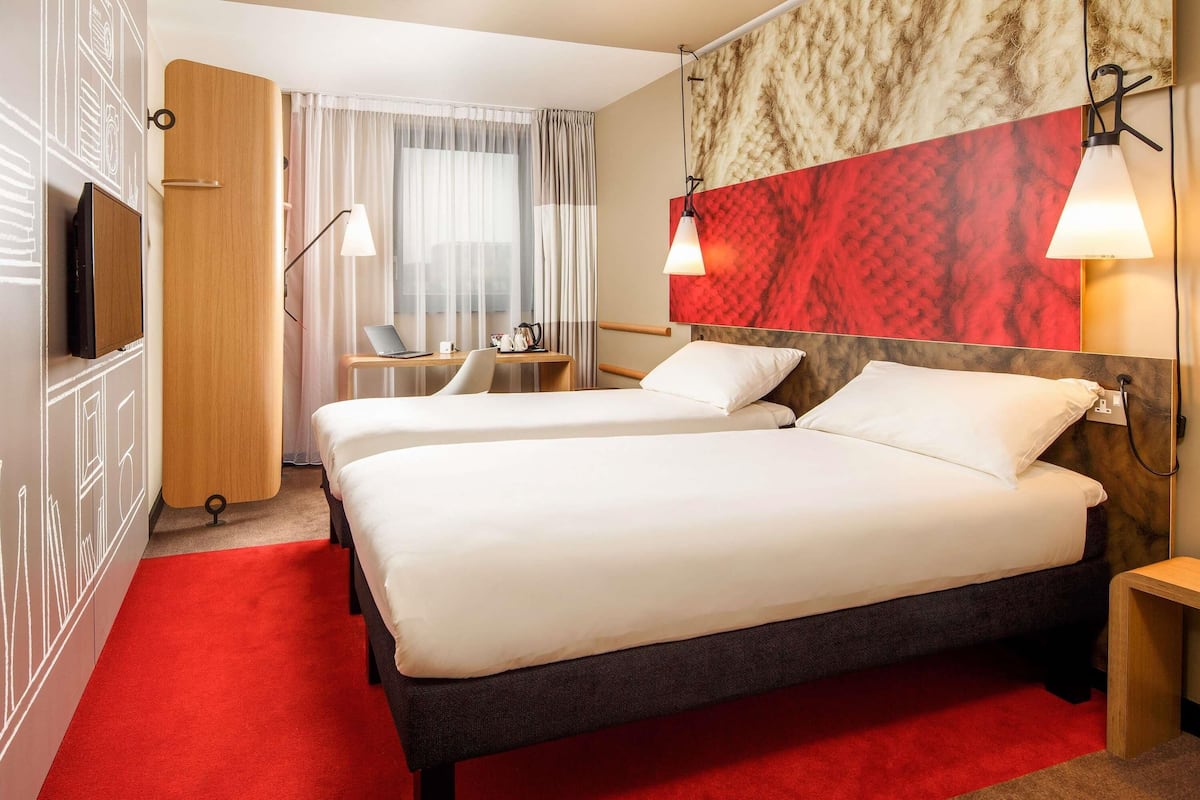 A hotel room with two beds, a desk with a laptop, a chair, and a red carpet.