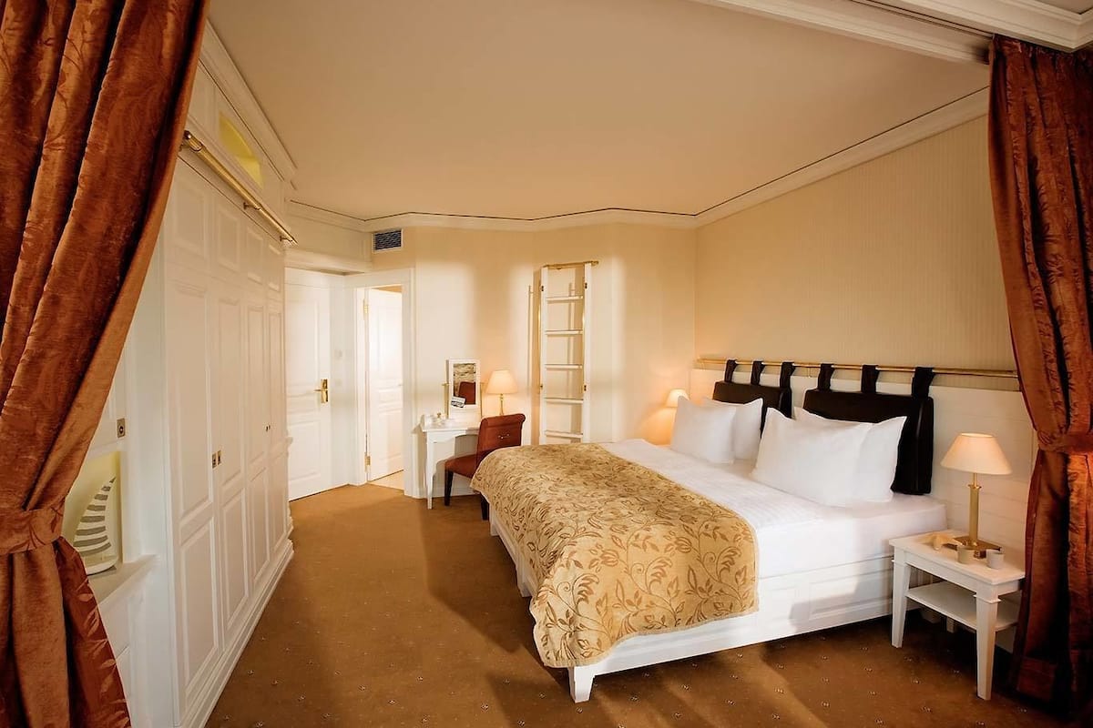 Ein Hotelzimmer mit einem großen Bett, einem Schreibtisch und einem Stuhl. Im Eck befindet sich ein Kleiderschrank und ein Spiegel.