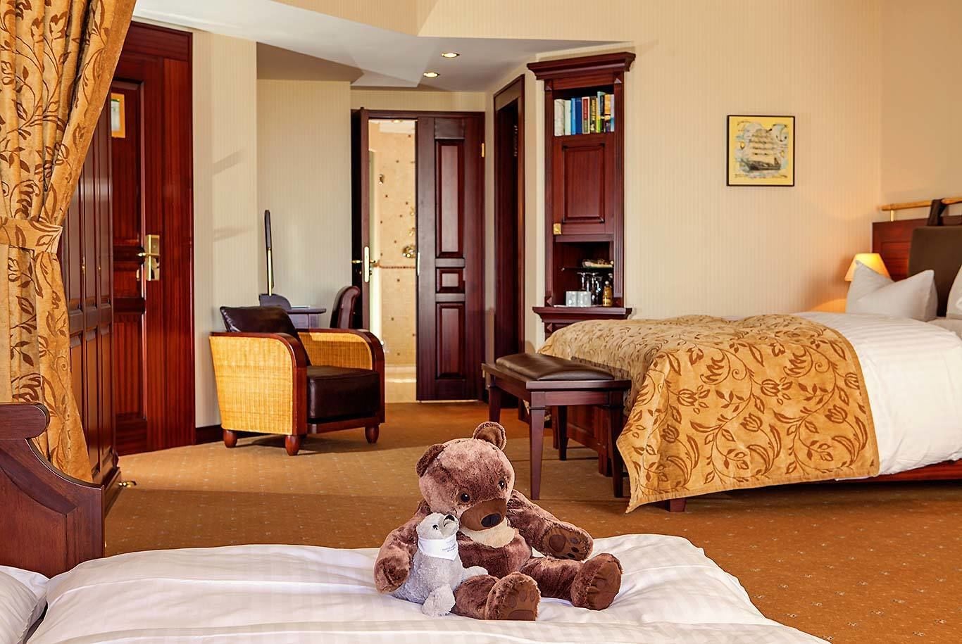 A hotel room with a bed, a chair, a desk, a bookshelf, and a teddy bear on the bed.