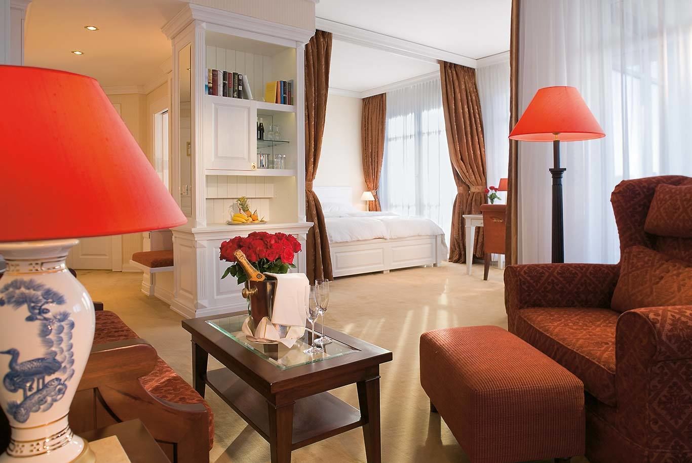 A hotel room with a bed, a seating area with a red armchair, a table with a vase of flowers, and a bookshelf.