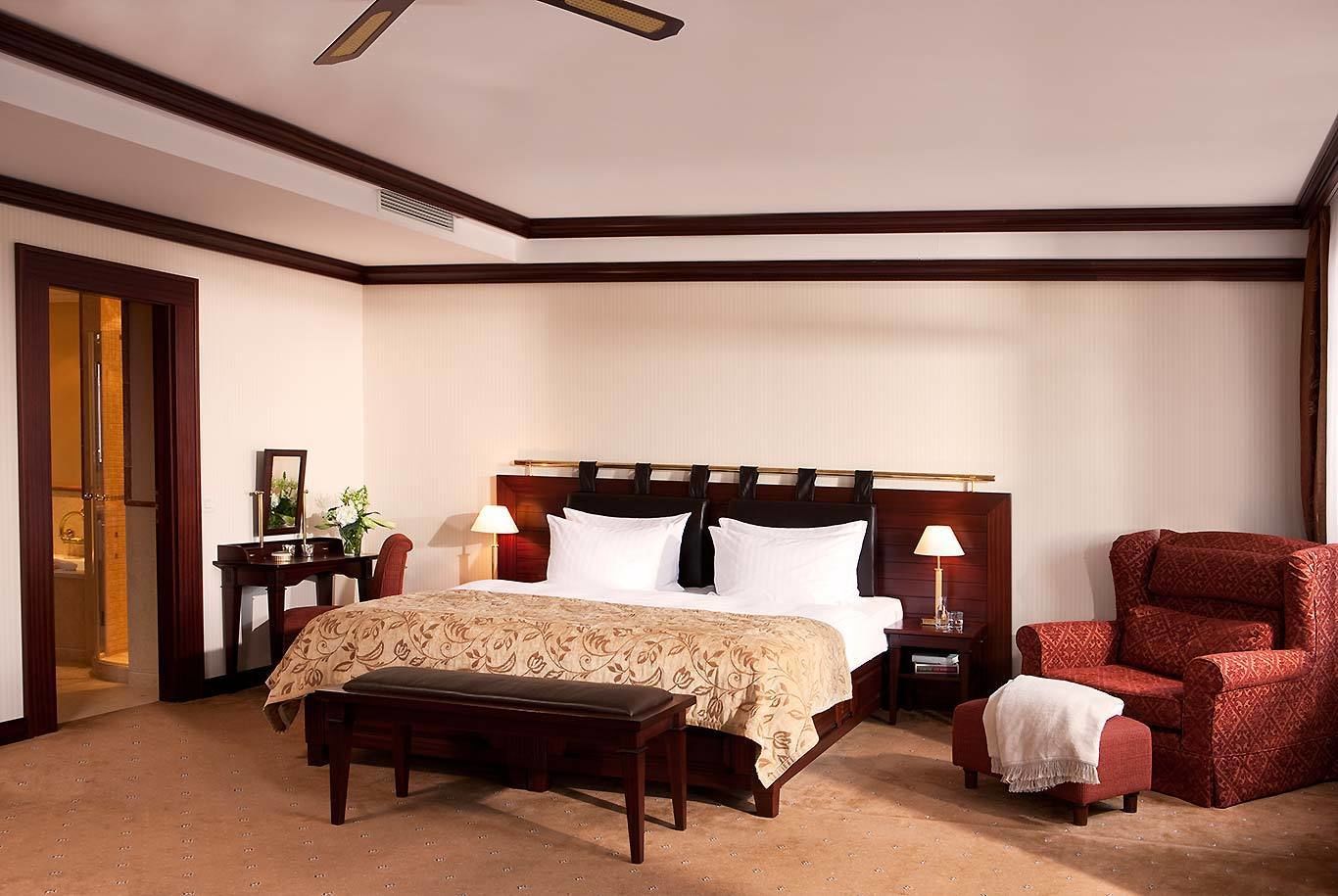 A hotel room with a large bed, a wooden headboard, a bench, a red armchair, and a nightstand with a lamp.