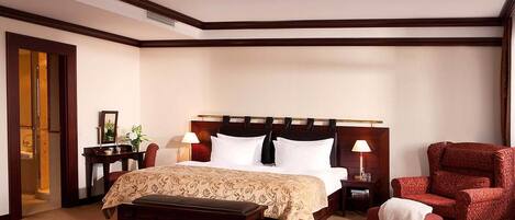 A hotel room with a large bed, a wooden headboard, a bench, a red armchair, and a nightstand with a lamp.