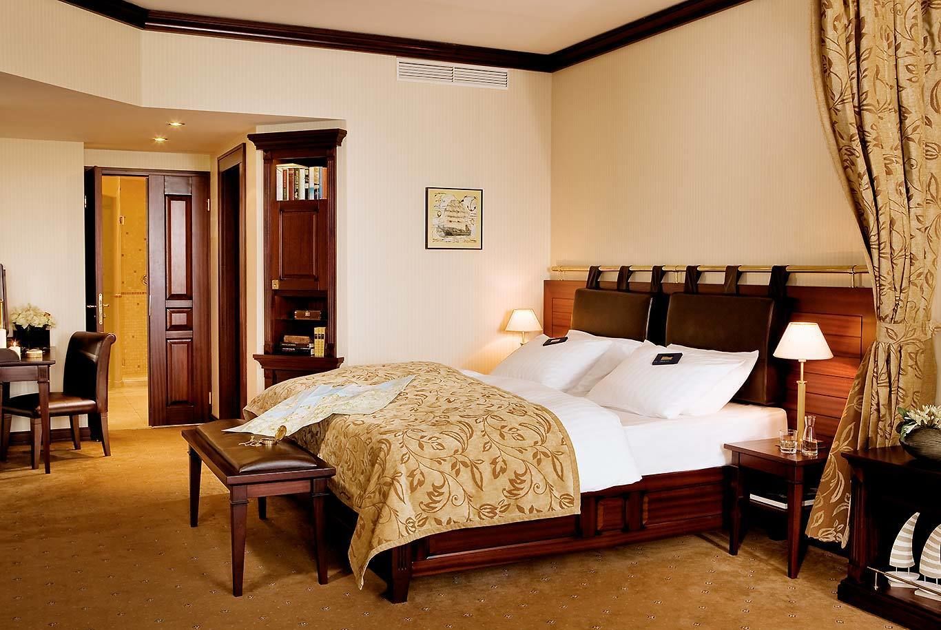 A hotel room with a large bed, a wooden headboard, a bedside table with a lamp, a desk with a chair, and a wardrobe.