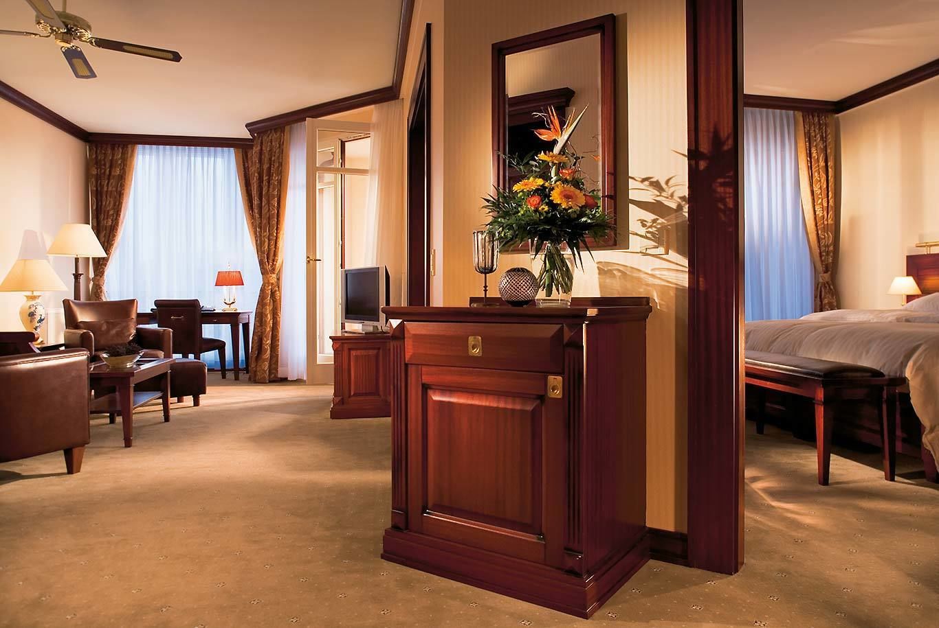 Une chambre d’hôtel avec un grand lit, un bureau avec une télévision, un coin salon avec des fauteuils et un grand miroir.