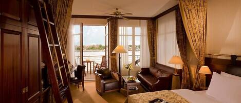A hotel room with a bed, a sofa, a desk with a lamp, a chair, a table with a vase of flowers, and a view of a body of water through large windows.