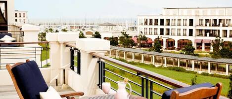 A balcony with two wooden chairs and a table, overlooking a marina with boats and a building with a restaurant.