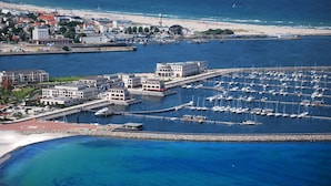 Eine Luftaufnahme einer Marina mit zahlreichen angestaubten Booten, einem Strand im Hintergrund und Gebäuden entlang der Uferlinie.