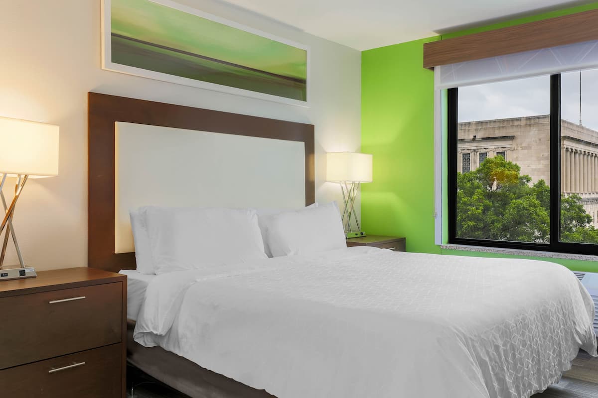 A hotel room with a bed, two bedside tables, a window with a view of a building, and a green wall.