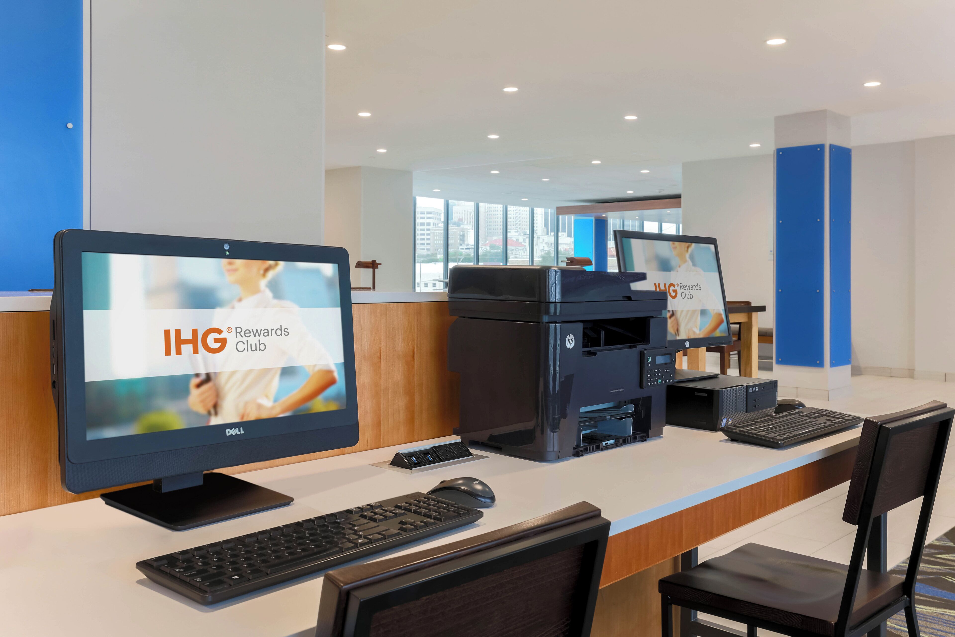 Una mesa de recepción con un monitor de computadora mostrando 'IHG Rewards Club', una impresora, un teclado y un ratón.
