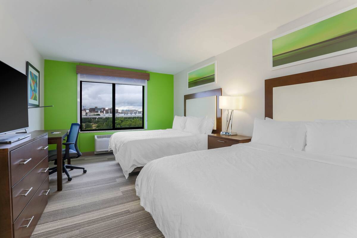 A hotel room with two beds, a desk, a chair, and a large window offering a city view.