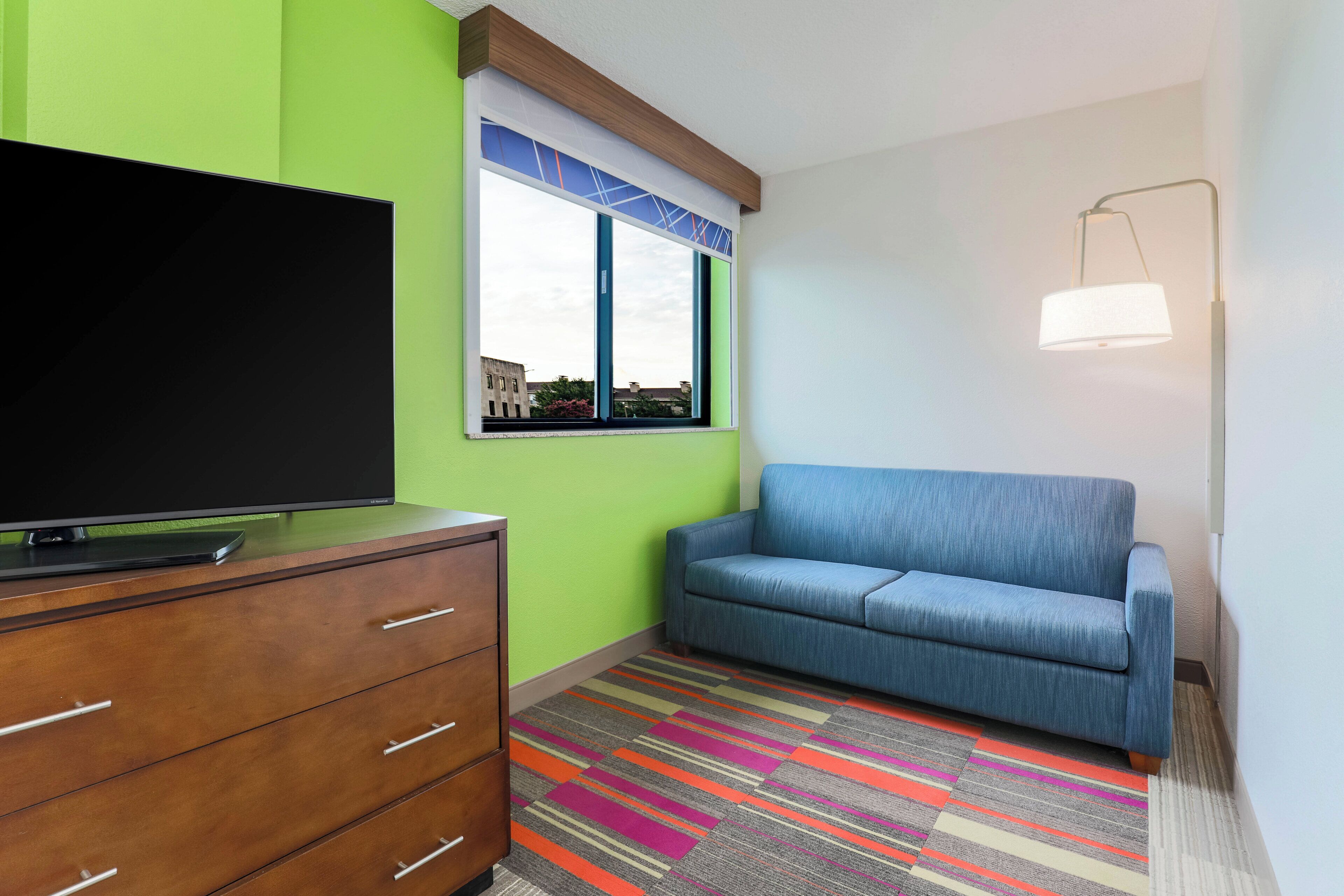 Une pièce avec un téléviseur à écran plat posé sur une commode en bois, un canapé bleu, un tapis rayé aux couleurs vives et une fenêtre donnant sur des immebles.