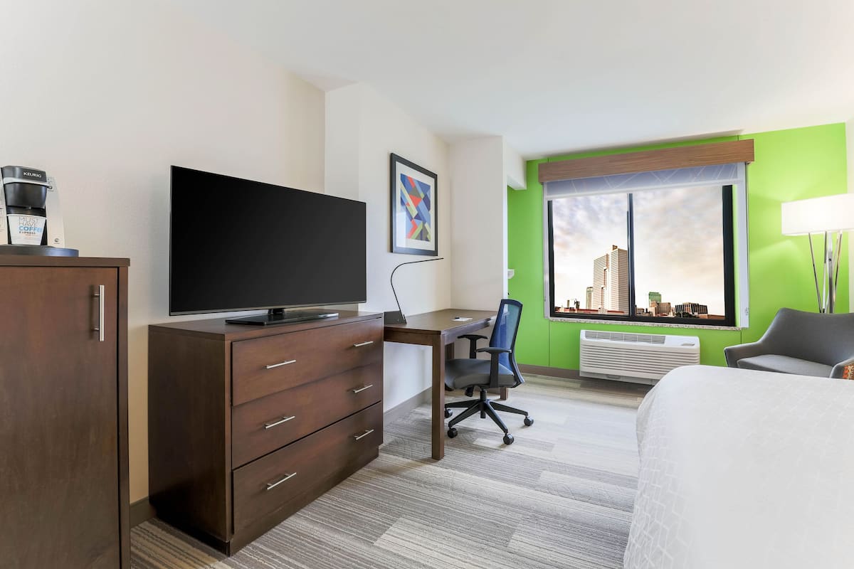 A hotel room with a flat-screen TV, a desk with a chair, a bed, a wardrobe, a framed picture, a window with a city view, and a lamp.