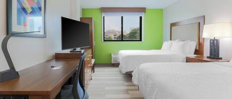 A hotel room with a desk, a television, two beds, a window, and a lamp.