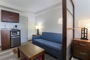 A compact hotel room with a kitchenette, a sofa, a wooden table, and a lamp. - Holiday Inn Express & Suites Fort Worth Downtown by IHG (Fort Worth)