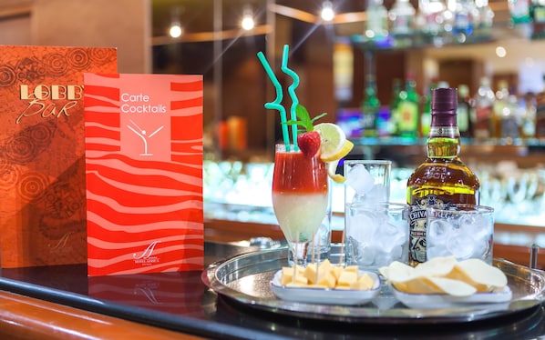Zona de bar con carta de cócteles, una bebida con pajita y una botella de Chivas Regal sobre una bandeja con queso y galletas.